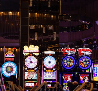 A lively casino scene featuring a row of bright, colorful slot machines with themes like Monopoly, Michael Jackson, and Lord of the Rings. The gaming machines display a variety of flashing lights and digital screens, creating an energetic and exciting atmosphere. Reflections of the lights are visible in glossy surfaces above the machines.