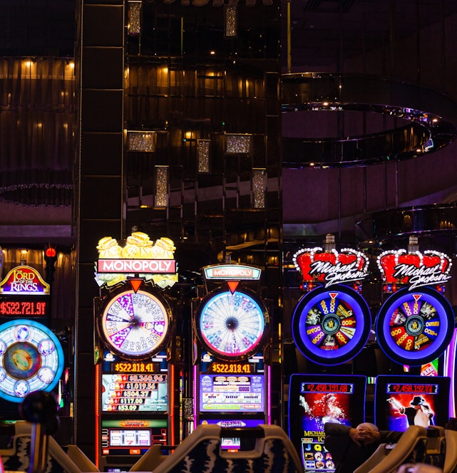 A lively casino scene featuring a row of bright, colorful slot machines with themes like Monopoly, Michael Jackson, and Lord of the Rings. The gaming machines display a variety of flashing lights and digital screens, creating an energetic and exciting atmosphere. Reflections of the lights are visible in glossy surfaces above the machines.