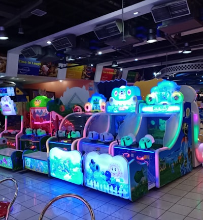 A brightly lit arcade filled with colorful gaming machines sits under ceiling lights. Neon lights illuminate various game stations, creating a playful and vibrant environment. A few red chairs are scattered around the reflective, tiled floor, and a person is observing one of the machines. The atmosphere is lively and cheerful, featuring games with cartoonish designs and engaging themes.